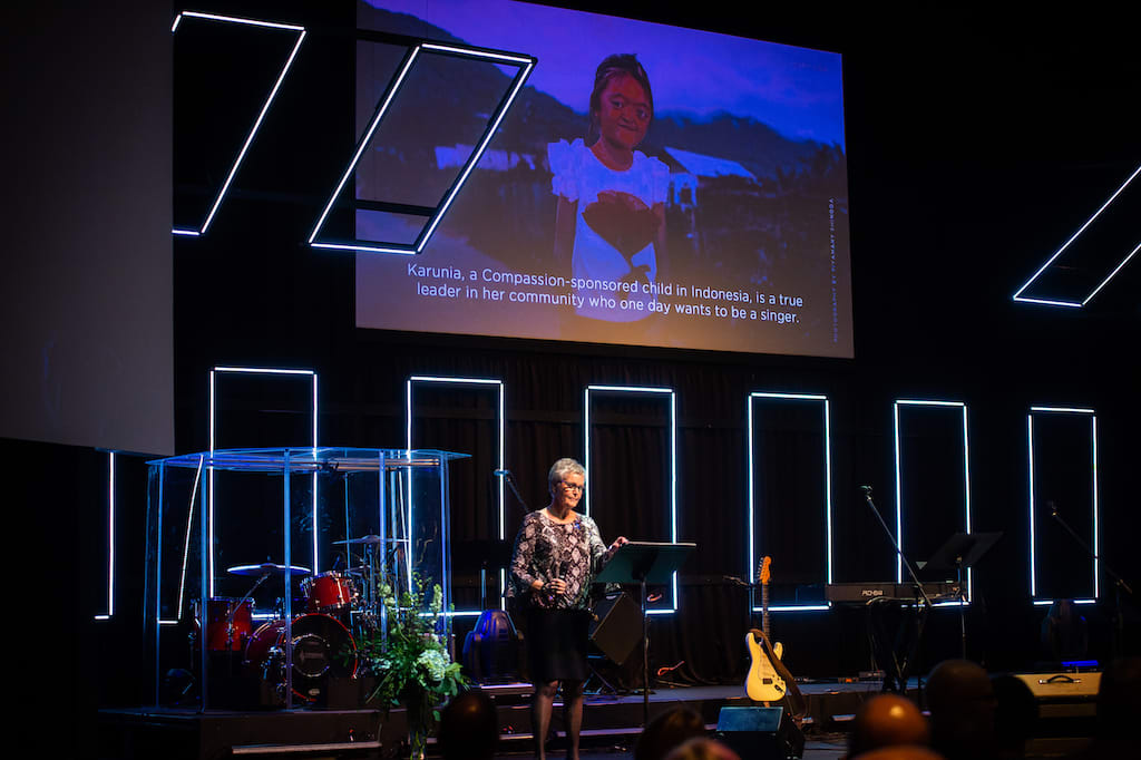 A woman stands on stage with the photo of a Compassion-sponsored child, Karunia, on the slide behind her.